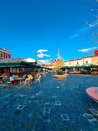 A vibrant outdoor caf&eacute; setting with various colorful chairs and tables under green canopies, set on a cobblestone plaza. The sky is clear and blue, with a few scattered clouds, surrounded by buildings with a mix of old-world charm and modern design. There are some trees with sparse leaves, indicating a mild season.