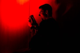 A person stands in a dimly lit environment, holding a camera or similar device. The scene is bathed in intense red light, creating a dramatic and mysterious atmosphere. Shadows and contrast are prominent, obscuring some details.