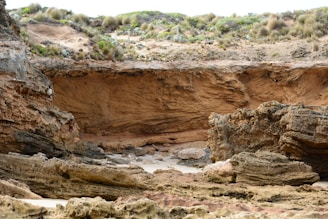 A rugged coastline with large, weathered rock formations and cliffs. The rough textures of the rocks contrast with patches of greenery and sandy areas. The scene is natural and untouched, showcasing erosion and diverse geological layers.