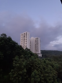 Two tall apartment buildings rise amidst a lush landscape of dense green trees, set against a backdrop of rolling hills and overcast skies.
