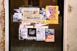 A collection of various posters and flyers covering a glass window. The posters include messages about environmental and social issues, with visible texts like 'Rifiuti Mondiali', 'Marcia per il Clima', and 'Nazieuropa'. The layout is eclectic with overlapping materials, and the colors are vibrant, especially the yellow and orange posters.