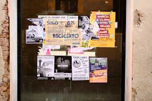 A collection of various posters and flyers covering a glass window. The posters include messages about environmental and social issues, with visible texts like 'Rifiuti Mondiali', 'Marcia per il Clima', and 'Nazieuropa'. The layout is eclectic with overlapping materials, and the colors are vibrant, especially the yellow and orange posters.