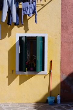 Clothing hangs on a line against a bright yellow wall, with a window featuring green shutters below it. A red broom leans against the wall next to a blue bucket, and a white bottle is placed on the ground.