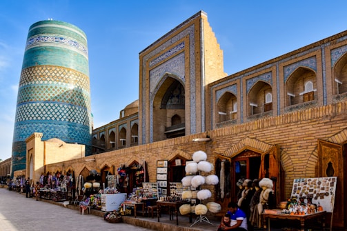 An ancient marketplace with traditional architecture featuring a prominent decorated blue cylindrical tower and archways with intricate designs. Various shops along the walkway display an array of culturally significant items and souvenirs, including hats and other local crafts. People are seen browsing and interacting at the market.