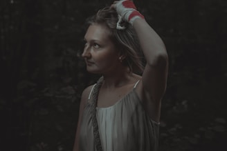 A woman with a contemplative expression stands in a dimly lit outdoor setting. She has a bandaged hand placed on her head and is wearing a sleeveless dress. The background suggests a forest or wooded area due to the dense, dark foliage.