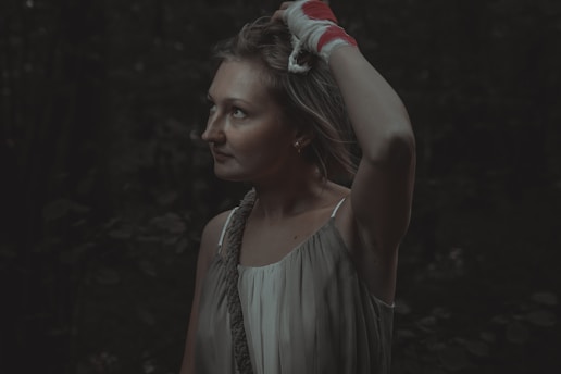 A woman with a contemplative expression stands in a dimly lit outdoor setting. She has a bandaged hand placed on her head and is wearing a sleeveless dress. The background suggests a forest or wooded area due to the dense, dark foliage.