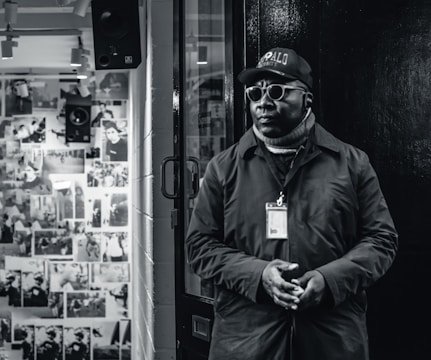 A person wearing a coat, scarf, and a cap stands near a doorway. They have sunglasses on and an identification badge around their neck. The background features a wall filled with photographs and a speaker.