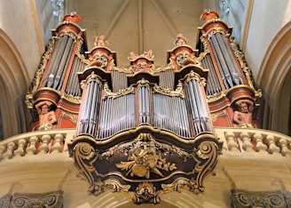 A grand and ornate pipe organ inside a cathedral, featuring elaborate decorative elements and sculptures of human figures integrated into its design. The organ pipes are framed by intricate carvings in gold and red hues, highlighting the craftsmanship and architectural detail.