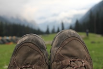 A pair of worn hiking boots is in the foreground, with a blurred background of a grassy field, distant mountains, and a cloudy sky. Small tents can be seen to the side, with trees lining the background.