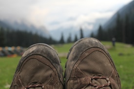 A pair of worn hiking boots is in the foreground, with a blurred background of a grassy field, distant mountains, and a cloudy sky. Small tents can be seen to the side, with trees lining the background.