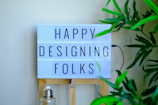 A lightbox sign displays the message 'Happy Designing Folks' in capital letters. The lightbox is placed on a wooden stand and surrounded by green plant leaves. There is a bottle with a metal cap in front of the stand, giving a casual and creative workspace vibe.