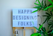 A lightbox sign displays the message 'Happy Designing Folks' in capital letters. The lightbox is placed on a wooden stand and surrounded by green plant leaves. There is a bottle with a metal cap in front of the stand, giving a casual and creative workspace vibe.