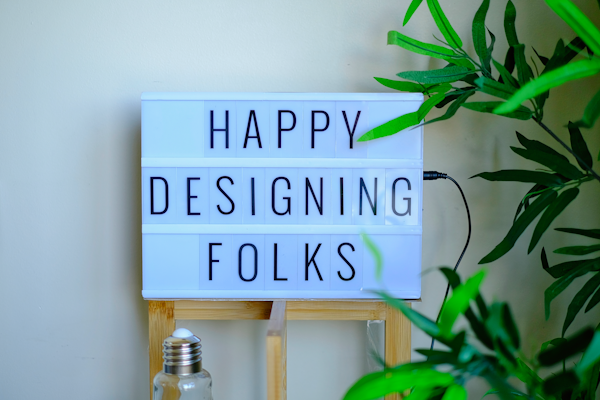A lightbox sign displays the message 'Happy Designing Folks' in capital letters. The lightbox is placed on a wooden stand and surrounded by green plant leaves. There is a bottle with a metal cap in front of the stand, giving a casual and creative workspace vibe.