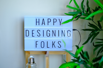 A lightbox sign displays the message 'Happy Designing Folks' in capital letters. The lightbox is placed on a wooden stand and surrounded by green plant leaves. There is a bottle with a metal cap in front of the stand, giving a casual and creative workspace vibe.