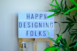 A lightbox sign displays the message 'Happy Designing Folks' in capital letters. The lightbox is placed on a wooden stand and surrounded by green plant leaves. There is a bottle with a metal cap in front of the stand, giving a casual and creative workspace vibe.