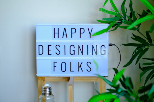 A lightbox sign displays the message 'Happy Designing Folks' in capital letters. The lightbox is placed on a wooden stand and surrounded by green plant leaves. There is a bottle with a metal cap in front of the stand, giving a casual and creative workspace vibe.