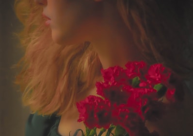 A softly lit, artistic photograph of a person's profile with long, wavy hair cascading down their shoulder. They are holding a bouquet of vibrant red carnations close to their chest. The overall lighting is warm, enhancing the soft texture of the hair and the vividness of the flowers.