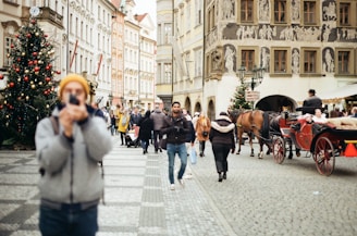 A busy street scene in a European city, featuring a group of people walking on a cobblestone path. Several of them are dressed in winter clothing. A person in the foreground is taking a photo, slightly out of focus. On the right side, a horse-drawn carriage with passengers goes by, and there are two decorated Christmas trees adding a festive atmosphere.