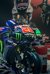 Several racing motorcycles are displayed with prominent logos, such as Monster Energy and Yamaha, on their bodies. Bold numbers like 20 and 99 are visible, with sleek designs indicating high performance. The motorcycles are positioned in a showroom setting with a polished floor and spotlights highlighting their aerodynamic shapes.