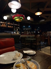 A dimly lit restaurant with an industrial design. There is a red leather seat in the foreground, with a table showing leftover food on plates and used napkins. In the background, there are several round tables and chairs. Shelves lining the upper part of the walls are stocked with various items and a counter is staffed by people. A colorful and artistic chandelier made from bottles hangs from the ceiling.