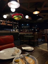 A dimly lit restaurant with an industrial design. There is a red leather seat in the foreground, with a table showing leftover food on plates and used napkins. In the background, there are several round tables and chairs. Shelves lining the upper part of the walls are stocked with various items and a counter is staffed by people. A colorful and artistic chandelier made from bottles hangs from the ceiling.