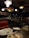 A dimly lit restaurant with an industrial design. There is a red leather seat in the foreground, with a table showing leftover food on plates and used napkins. In the background, there are several round tables and chairs. Shelves lining the upper part of the walls are stocked with various items and a counter is staffed by people. A colorful and artistic chandelier made from bottles hangs from the ceiling.