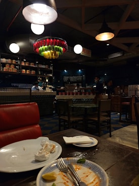 A dimly lit restaurant with an industrial design. There is a red leather seat in the foreground, with a table showing leftover food on plates and used napkins. In the background, there are several round tables and chairs. Shelves lining the upper part of the walls are stocked with various items and a counter is staffed by people. A colorful and artistic chandelier made from bottles hangs from the ceiling.