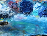 A vibrant and abstract mix of oil colors is smeared and blended on a palette surface, featuring a spectrum of blues, whites, yellows, and reds with thick, textured strokes.