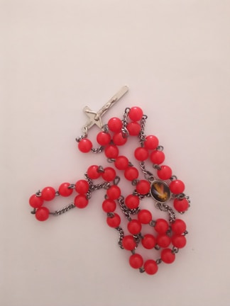 A rosary with red beads is laid out on a light surface. It features a silver crucifix and a small medallion attached with metal chain links. The beads are uniformly round and vividly colored.
