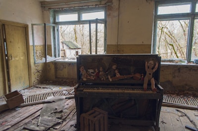 An abandoned room with peeling paint and broken windows. A neglected piano stands in the center, with several old dolls placed on top of it. Leaves and debris are scattered on the wooden floor, and trees are visible through the windows.