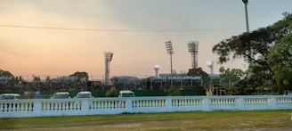 A sports stadium with tall floodlights and a seating area filled with people wearing white shirts. Several vehicles are parked in front. The scene is framed by trees with a warm, late afternoon sky.