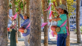 People are participating in an outdoor activity, each holding a colorful pink ribbon attached to a stick and wearing drums tied around their waists. They are surrounded by several trees in a park setting, wearing casual clothing and sun hats.