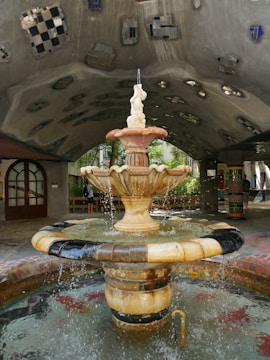A textured fountain with an ornate sculpture at the top, surrounded by a pool of water with visible ripples and splashes. The area is covered by a unique arched concrete ceiling, adorned with small, abstract tile patterns. Natural light filters through greenery in the background, creating a serene ambiance.