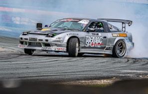 A high-performance race car is captured performing a drift maneuver on a race track. The car has multiple sponsor logos, a prominent rear wing, and low-profile tires. Smoke trails from the rear wheels, indicating the car is in motion. The background includes a blurred race track barrier, suggesting high speed.