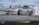 A high-performance race car is captured performing a drift maneuver on a race track. The car has multiple sponsor logos, a prominent rear wing, and low-profile tires. Smoke trails from the rear wheels, indicating the car is in motion. The background includes a blurred race track barrier, suggesting high speed.
