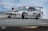 A high-performance race car is captured performing a drift maneuver on a race track. The car has multiple sponsor logos, a prominent rear wing, and low-profile tires. Smoke trails from the rear wheels, indicating the car is in motion. The background includes a blurred race track barrier, suggesting high speed.