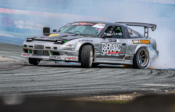 A high-performance race car is captured performing a drift maneuver on a race track. The car has multiple sponsor logos, a prominent rear wing, and low-profile tires. Smoke trails from the rear wheels, indicating the car is in motion. The background includes a blurred race track barrier, suggesting high speed.