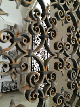 Intricate wrought iron patterns dominate the foreground, with details of floral and swirling designs. In the background, there are black-and-white photographs displayed on a white wall, giving a historical or artistic feel to the setting.