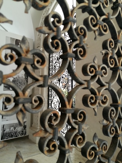 Intricate wrought iron patterns dominate the foreground, with details of floral and swirling designs. In the background, there are black-and-white photographs displayed on a white wall, giving a historical or artistic feel to the setting.