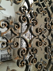 Intricate wrought iron patterns dominate the foreground, with details of floral and swirling designs. In the background, there are black-and-white photographs displayed on a white wall, giving a historical or artistic feel to the setting.