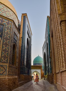 A narrow, cobblestone passageway flanked by beautifully ornate and colorful tiled buildings leads towards a domed structure. The tiles display intricate geometric patterns in shades of blue and green. A person in a vibrant dress walks down the path, adding a touch of liveliness to the serene architectural setting.