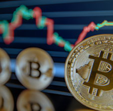 Three golden bitcoins are positioned in front of a fluctuating line graph displaying financial data with red and green lines against a dark background. The image conveys a focus on cryptocurrency and market trends.