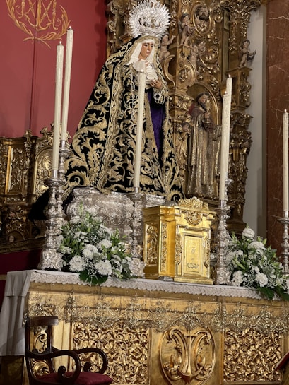 A detailed altar featuring an ornate religious statue draped in a black and gold robe with an intricate design. The statue has a crown and is holding a heart, standing amidst tall, white candles. Flowers and greenery are arranged around the base, and the altar is decorated with rich gold detailing, including a golden tabernacle. The background has a deep red wall with a gold emblem and elaborate carvings.