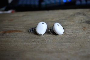Two small white wireless earbuds are placed on a wooden surface. These earbuds have a sleek design with a metallic accent and are positioned with the protective tips facing outward. The background is slightly blurred, creating a soft focus effect.