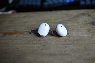 Two small white wireless earbuds are placed on a wooden surface. These earbuds have a sleek design with a metallic accent and are positioned with the protective tips facing outward. The background is slightly blurred, creating a soft focus effect.