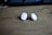 Two small white wireless earbuds are placed on a wooden surface. These earbuds have a sleek design with a metallic accent and are positioned with the protective tips facing outward. The background is slightly blurred, creating a soft focus effect.