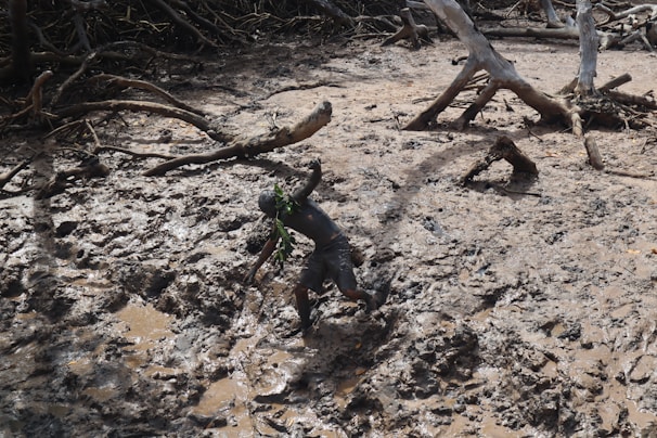 A person is struggling to walk through a dense, muddy terrain, surrounded by scattered tree branches and roots. The environment looks rugged and untamed, as the person uses hands for balance.
