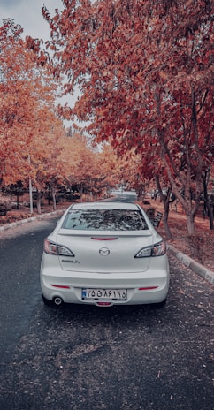 A white Mazda 3 is parked on a tree-lined road, surrounded by autumn foliage with vibrant orange and red leaves.