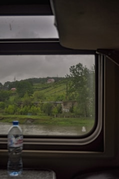 A view through a train window reveals a rural landscape with lush green hills, a river, and a distant house set amidst trees. A bottle is placed on the window sill inside the train.