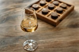 A glass of white wine with 'Sycamore' branding sits on a wooden surface. In the background, there is a wooden board game, possibly tic-tac-toe, with round game pieces.
